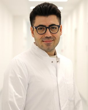 Ein Arzt in einem weißen Kittel steht in einem hellen Flur und lächelt, mit Brille und gepflegtem Aussehen.