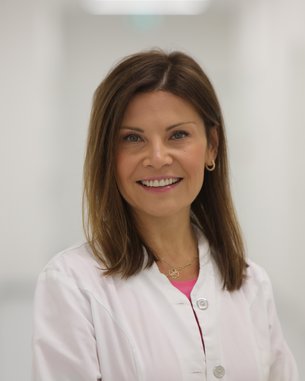 Eine lächelnde Frau in einem weißen Kittel steht in einem hellen, modernen Flur. Sie hat lange, braune Haare und trägt goldene Ohrringe.