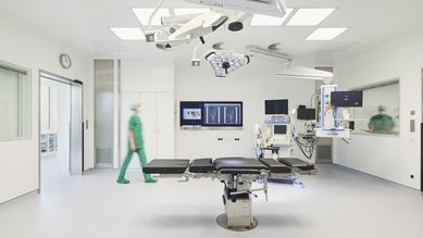 Operationssaal mit einem chirurgischen Tisch in der Mitte. Ein medizinisches Gerät steht neben dem Tisch, während eine Person in grüner Kleidung den Raum betritt. An den Wänden sind Monitore und medizinische Ausrüstung sichtbar.