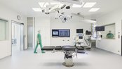 Operationssaal mit einem chirurgischen Tisch in der Mitte. Ein medizinisches Gerät steht neben dem Tisch, während eine Person in grüner Kleidung den Raum betritt. An den Wänden sind Monitore und medizinische Ausrüstung sichtbar.